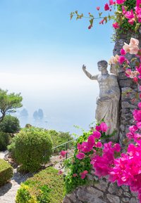 Steinstatue mit erhobenem Arm vor einem Hintergrund aus lebhaftem pinken Bougainvillea, grünem Laub und einem Küstenblick auf entfernte Felsen und das Meer.
