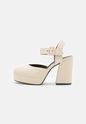 Zapato de tacón alto con plataforma beige, correa en el tobillo con hebilla, punta redondeada y tacón ancho en bloque sobre fondo blanco.