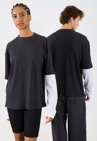 Czarna, żeberkowana koszulka z krótkim rękawem z białymi długimi rękawami pod spodem. Koszulka ma luźny krój, okrągły dekolt i gładką teksturę.