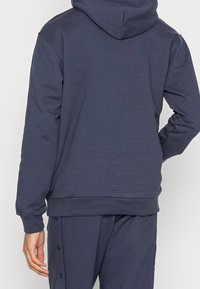 Person med mörkblå huvtröja och matchande byxor, sedd bakifrån med en hand vilande på höften.