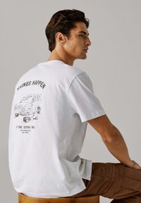 Wit katoenen T-shirt met korte mouwen, met een grafische print van een busje en tekst op de achterkant. Model getoond in profiel, zittend.