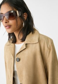 Tan ruskindsjakke med krave og en enkelt sort knap. Model iført oversize brune solbriller og en hvid ribbet top.