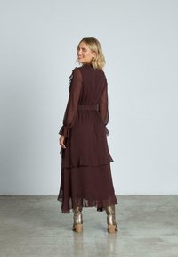 Lång bordeauxfärgad klänning med volangdetaljer, genomskinliga ärmar och åtdragen midja. Kombinerad med glänsande guldiga ankelstövlar.