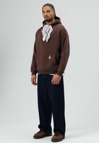 Bruine hoodie met een grote voorkantzak, voorzien van een witte bandana gestrikt rond de nek en een klein logo in de hoek. Donkere, loszittende jeans en bruine laarzen maken de look compleet.