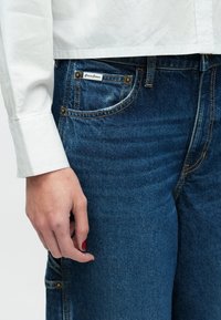 Ξεθωριασμένο μπλε τζιν με άνετη γραμμή, χρυσά μεταλλικά στοιχεία, ορατή ραφή και λευκή ετικέτα στην τσέπη. Μερικώς ορατό λευκό κομμένο τοπ.