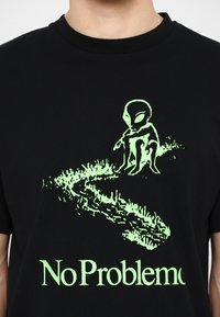 Sort bomulds T-shirt med et lysende grønt alien-illustration og teksten "No Problemo" under, hvilket skaber et iøjnefaldende grafisk design.