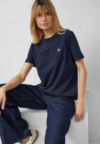 Frau mit schulterlangem blondem Haar, die ein marineblaues T-Shirt mit kleinem Herzlogo und lockere dunkle Jeans trägt und auf einem weißen Block sitzt.