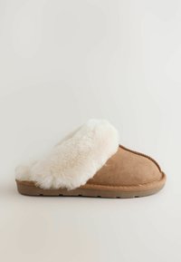 Beige mokasīni ar balto pūkaino oderi un teksturētu zoli. Sānu skats izceļ atvērtu aizmuguri un noapaļotu pirkstu dizainu.