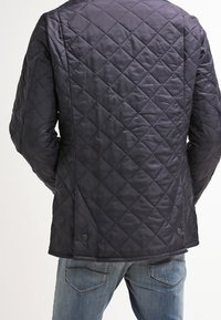 Schwarze gesteppte Jacke mit Rautenmuster, getragen von einer Person in blauen Jeans, von hinten betrachtet.