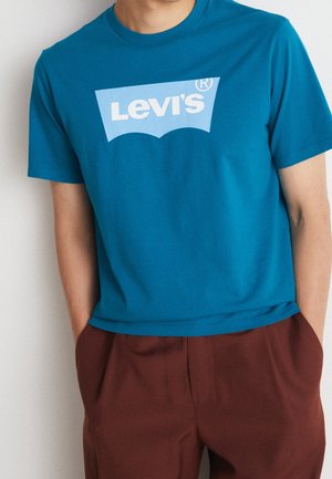 Hombre con camiseta verde azulado con el logo de Levi's metida dentro de pantalones marrones de cintura alta, con las manos en los bolsillos, frente a una pared blanca lisa.