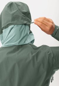 Giacca leggera con cappuccio in verde scuro e blu chiaro. Presenta un cordoncino sul retro del cappuccio per una vestibilità regolabile. Tessuto texturizzato.