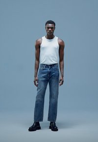 Top sin mangas blanco, jeans rectos azules y botas negras hasta el tobillo. Los jeans tienen un lavado desgastado y un diseño clásico de cinco bolsillos.