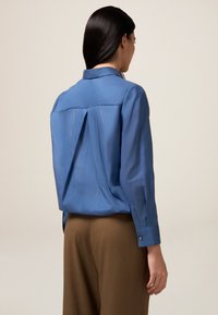Blaue Bluse mit langen Ärmeln, Rückenfalten, glatter Textur und einem Kragen. Kombiniert mit braunen Hosen, von hinten gesehen.