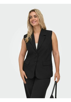 Femme souriante aux cheveux blonds portant un blazer noir sans manches et un pantalon noir, tenant un sac à main noir à côté d’elle.