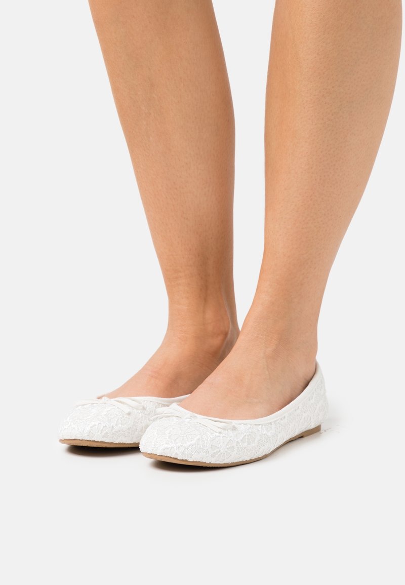 Pieds portant des ballerines en dentelle blanche avec de petits nœuds décoratifs sur un fond clair.