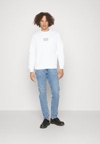 Felpa bianca con un piccolo logo grigio sul petto, abbinata a jeans azzurri chiari e scarpe nere, caratterizzata da una vestibilità casual e maniche lunghe.