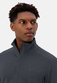 Giacca grigia con zip, in tessuto testurizzato e colletto alto. Presenta un logo discreto sul petto, progettata per il comfort e le attività all'aperto.