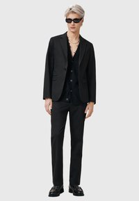 Zwarte suit met een op maat gemaakte jas en strakke broek, gecombineerd met een zwart button-up shirt en stijlvolle zonnebril. Gelaagde stof en moderne pasvorm.