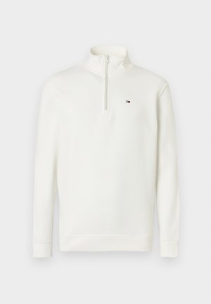 Baltas vilnos megztinis su aukštu apykakliu ir ketvirčio ilgio užtrauktuku. Ant krūtinės yra mažas logotipas, rankogaliai ir apačia yra ribrai. Minkšta tekstūra.