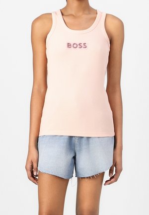 Frau trägt ein hellrosa Tanktop mit dem Schriftzug "BOSS" und hellblaue Denim-Shorts mit ausgefransten Kanten und steht vor einem weißen Hintergrund.