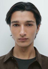 Młody mężczyzna o średniej długości ciemnych włosach, brązowych oczach i neutralnym wyrazie twarzy, ubrany w ciemną koszulę i brązową kurtkę na jednolitym tle.