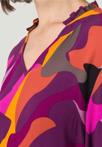 Blouse colorée avec des motifs de vagues abstraites en violet, orange, rose et gris. Elle a un col en V avec un bord volanté.