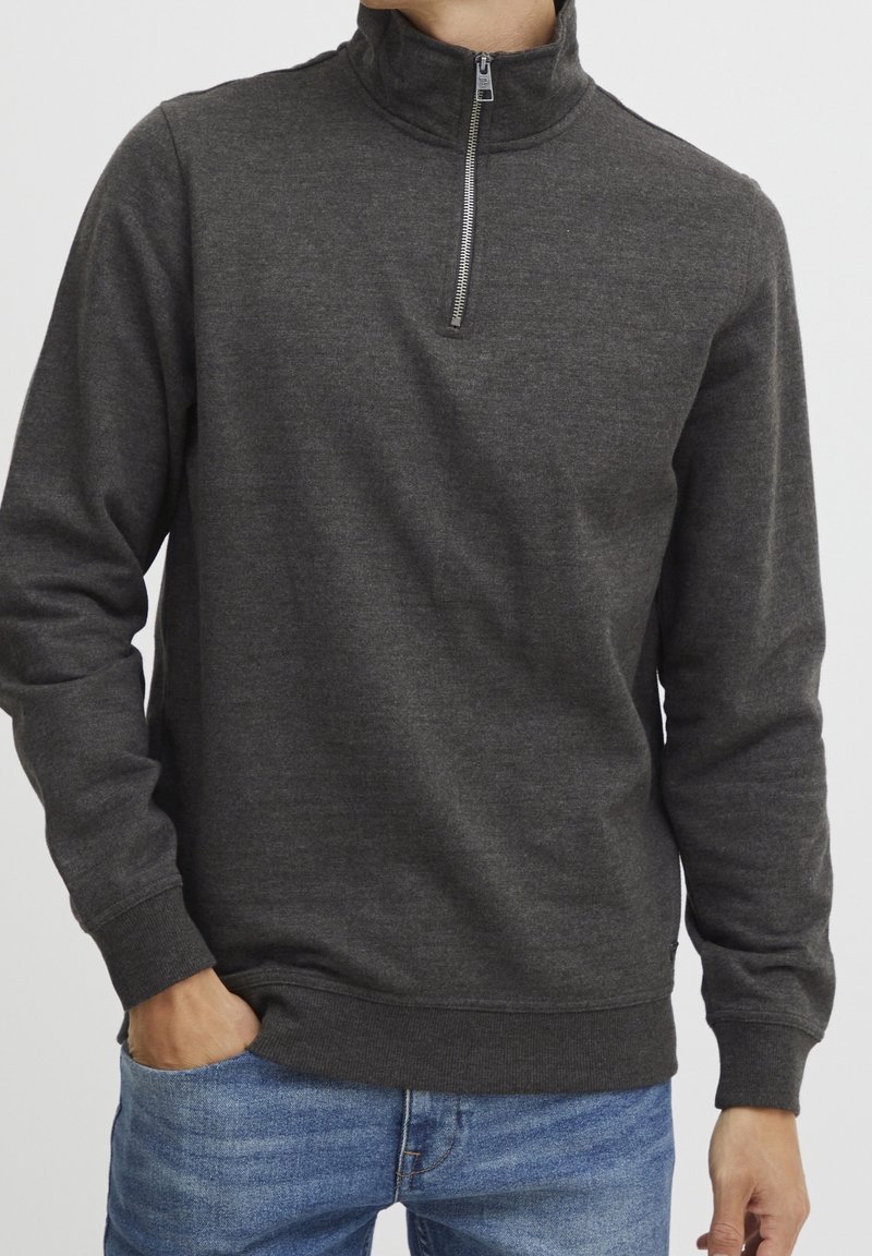Homme portant un pull gris foncé à col zippé et un jean bleu clair, main dans la poche, sur un fond uni.