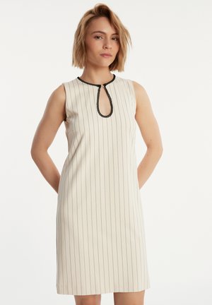 Mouwloze crèmekleurige jurk met zwarte verticale strepen, een keyhole halslijn afgewerkt met zwarte bies, en een rechte silhouette.