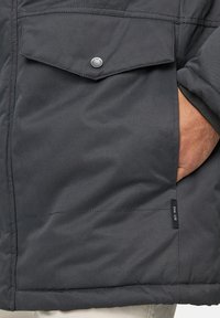 Chaqueta aislante de color gris oscuro que presenta un bolsillo con tapa y botón a presión, un bolsillo lateral y detalles de costura a lo largo de las costuras.