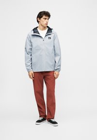 Νεαρός άνδρας που στέκεται, φορά ανοιχτό γκρι μπουφάν με φερμουάρ και κουκούλα, παντελόνι καφέ-σκουριάς και μαύρα αθλητικά παπούτσια, σε απλό λευκό φόντο.