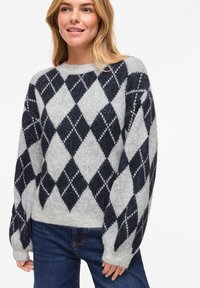 Pull en maille gris avec un motif argyle noir et blanc, col rond et manches surdimensionnées, porté avec un jean bleu.