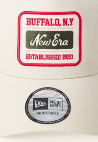 Gorra de béisbol de tela crema con un logo bordado que dice "Buffalo, N.Y." y "New Era", y una correa ajustable. Etiqueta negra en el borde.