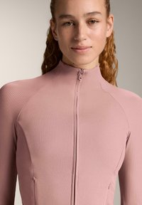 Fitted long-sleeve top in soft pink, featuring ribbed texture, high collar, and a front zipper. Two side pockets near the waist.