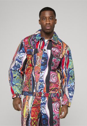 Hombre con una chaqueta de estampado colorido y pantalones a juego con diseños abstractos en rojo, azul, verde y negro, de pie frente a un fondo liso.