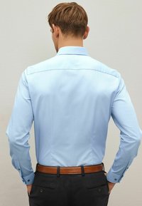 Hellblaues Button-up-Hemd mit langen Ärmeln, aus glattem Stoff, mit Rückenpasse und kontrastierenden Knöpfen an den Manschetten. Brauner Gürtel sichtbar.