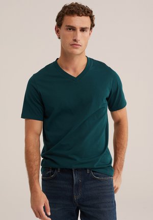 Jeune homme aux cheveux bouclés portant un t-shirt vert foncé à col en V à manches courtes et un jean bleu foncé sur un fond clair uni.