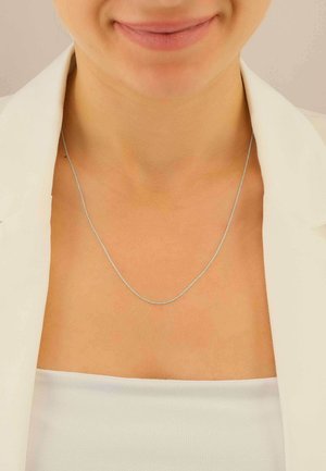Collana a catena d'argento con un design delicato e intrecciato, appoggiata sulla pelle nuda, su una maglia chiara e un blazer bianco.