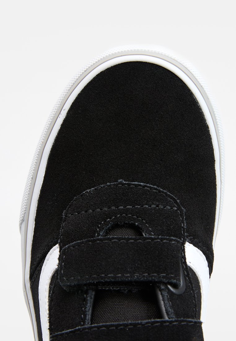 Zapatilla deportiva de ante negro con una franja blanca, con tiras de velcro y una parte superior texturizada. Punta redondeada y suela de goma.