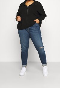 Svart stickad pullover med halvlång dragkedja i kragen, kombinerad med ljusblå trasiga jeans och vita sneakers. Avslappnad, ledig passform.