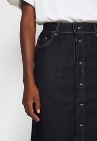 Tmavá džínsová sukně se žlutým prošíváním, předními knoflíky a bočními kapsami. Materiál má hladkou texturu a rovný střih.