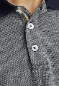 Pilkos tekstūros polo marškiniai su tamsiai mėlynu apykakle, trimis baltais mygtukais su tamsiomis siūlėmis ir kontrastinga vidine apykakle.