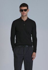Polo noir à manches longues côtelées avec un col, deux boutons, et un pantalon gris. Modèle portant des lunettes de soleil sombres, les mains dans les poches.