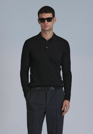 Black ribbed long-sleeve polo shirt with a collar, two buttons, and grey trousers. Model wearing dark sunglasses, hands in pockets.