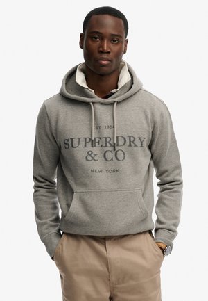 Mand iført en grå Superdry & Co hættetrøje over en skjorte med krave, beige bukser og et brunt ur, stående med hænderne i lommerne.