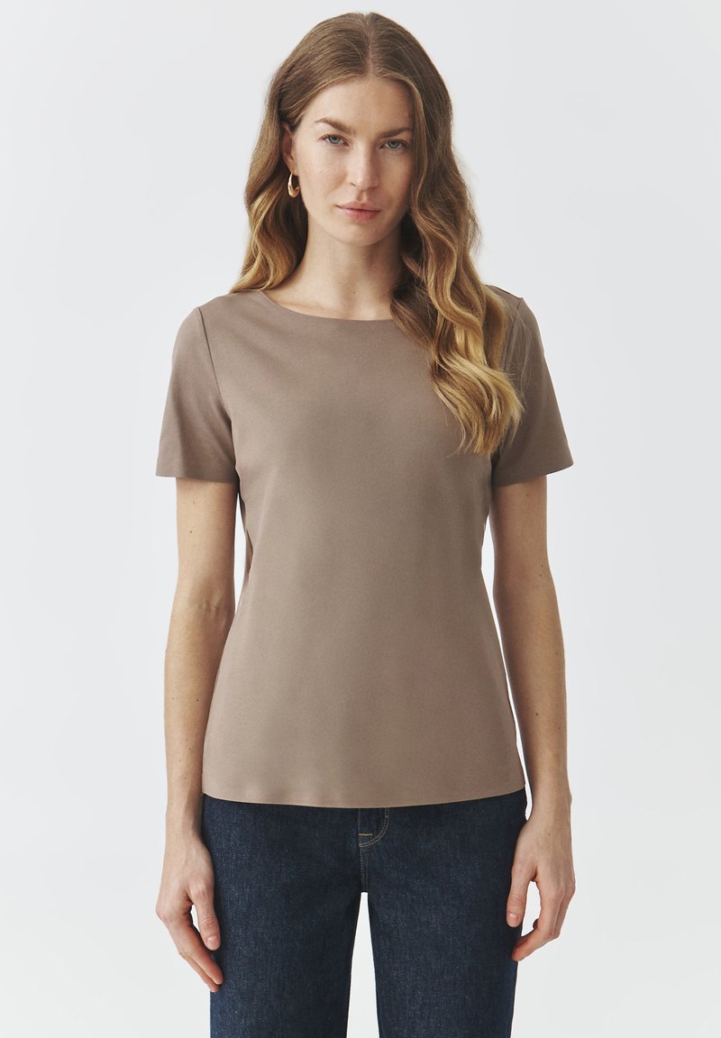 Top a maniche corte di un morbido color taupe, con scollatura rotonda e una texture liscia. Abbinato a jeans in denim scuro.