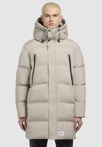 Chaqueta acolchada beige con un cuello alto, capucha ajustable con cordón, bolsillos laterales con cremallera y un acabado suave. Presenta un parche con el logo.