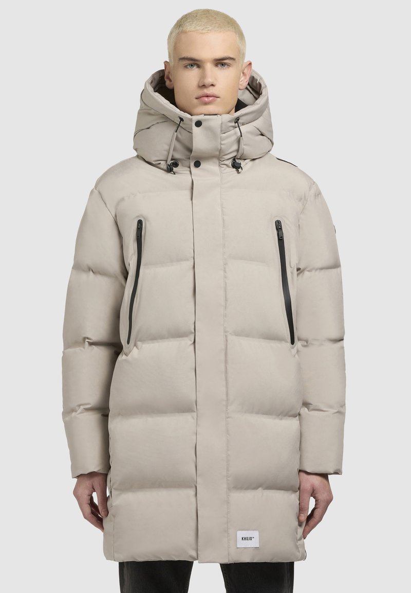 Chaqueta acolchada beige con un cuello alto, capucha ajustable con cordón, bolsillos laterales con cremallera y un acabado suave. Presenta un parche con el logo.