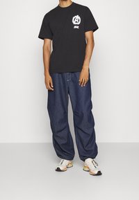 Černé bavlněné tričko s bílým grafickým logem na hrudi, kombinované s volnými modrými džíny a multikolorovými teniskami.