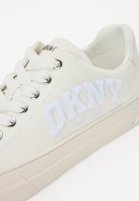 Fehér vászon tornacipő textúrált hímzett "DKNY" logóval, kerek orral, fém szemcsékkel és lapos fehér fűzőkkel, krémszínből készült talpon.