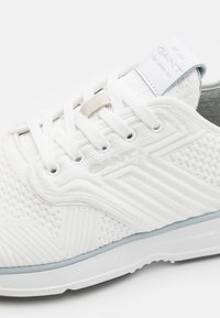 Baskets blanches GANT en tissu tricoté texturé, lacets blancs, logo discret sur le côté, et étiquette de languette de marque affichant les détails de la création.